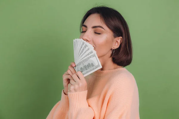 Young woman in casual peach sweater  isolated on green olive background  lucky holding fan of 100 dollar bills funny smell cash money copy space