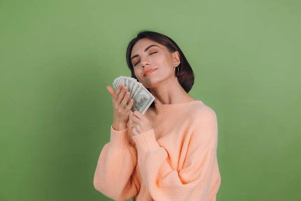 Young woman in casual peach sweater  isolated on green olive background  lucky holding fan of 100 dollar bills funny smell cash money copy space
