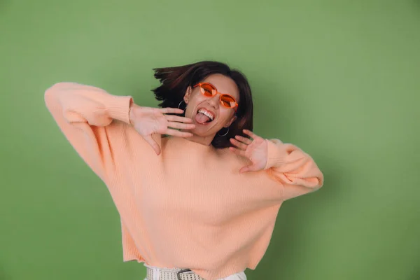 Young stylish woman in casual peach sweater and orange glasses isolated on green olive background happy positive funny jumping around copy space