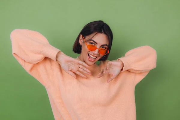 Young stylish woman in casual peach sweater and orange glasses isolated on green olive background happy positive smiling laugh around copy space