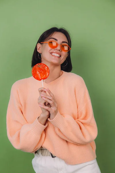 Young stylish woman in casual peach sweater and orange glasses isolated on green olive background with orange lollipop positive smile copy space