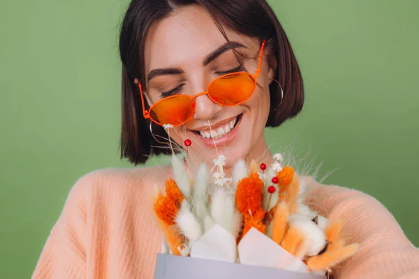 Young woman in casual peach sweater  isolated on green olive background  hold  orange white flower box composition of cotton flowers, gypsophila, wheat and lagurus for a gift happy surprised