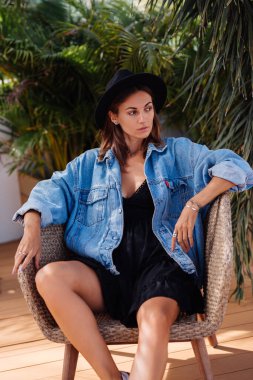 Beyaz elbiseli, şapkalı ve kot ceketli şık bir kadın. Sandalyeye otur, tropikal arka plan.