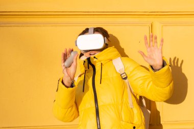 Sokakta sanal gerçeklik gözlüğü takan bir kadının portresi. Sarı bir duvara yaslanmış. Kışın parlak güneş ışığında, kalın giysiler içinde.