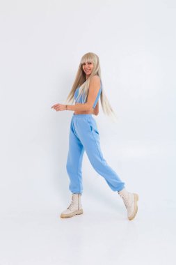 Beyaz arka planda, stüdyoda uzun, düz saçlı ve kâküllü mutlu bir sarışının moda portresi. Kız modaya uygun mavi bir takım elbise, kalın bir pantolon ve bir bluz giyiyor.