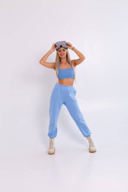 Stüdyo fotoğrafında, beyaz bir arka planda, sıcak mavi giysili, sanal gerçeklik gözlüğü takan çekici bir kadın var.
