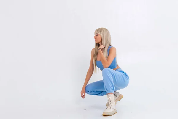 Fashion portrait of young happy blonde woman with long gorgeous straight hair and bangs in the studio on a white background. The girl is dressed in a trendy blue suit, warm pants and a top