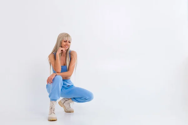 Fashion portrait of young happy blonde woman with long gorgeous straight hair and bangs in the studio on a white background. The girl is dressed in a trendy blue suit, warm pants and a top
