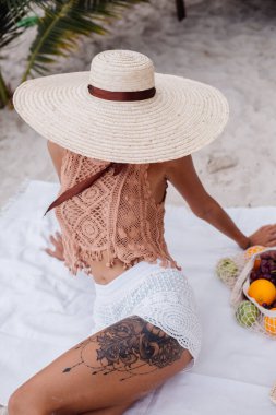 Kadın plaj halısı battaniyesinde oturmuş meyve sepetiyle piknik yapıyor. 