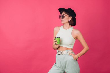 Şapkalı ve güneş gözlüklü güzel bir kadın pembe kırmızı arka planda poz veriyor. Elinde bir fincan kağıt kahve, izole edilmiş fotokopi alanı var.