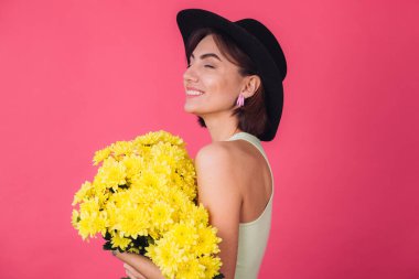 Pembe arka planda şapkalı şık bir kadın büyük bir buket sarı astar sarılıyor, bahar havasında, sakin bir şekilde gülümsüyor.