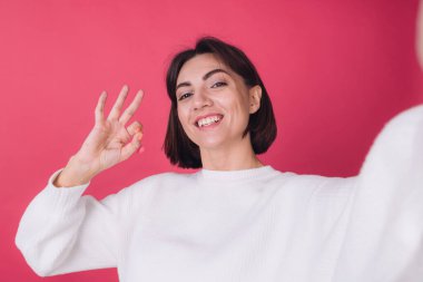 Gayri resmi beyaz süveter giyen güzel bir kadın pembe arka planda gülümseyen selfie çeken, parmaklarıyla işaret eden, izole edilmiş fotokopi alanını arayan bir video var.