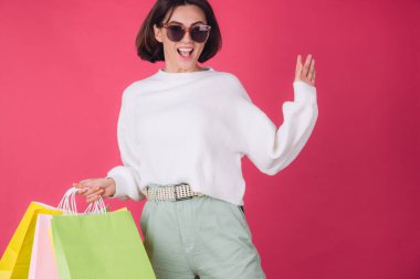Beyaz süveter ve pembe kırmızı arka planda güneş gözlüklü bir kadın alışveriş çantalarını tutar mutlu zıplar, heyecanlı dans eder izole edilmiş fotokopi alanı.