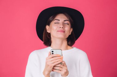 Beyaz süveter ve kırmızı arka planda şapka giyen şık bir kadın. Fotokopi uzay gülüşü, kapalı gözler, cep telefonu.