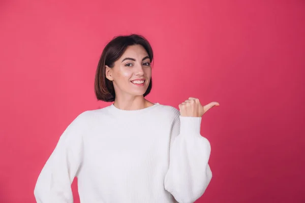 stock image Beautiful woman in casual white sweater on pink red background smiling point right with thumb finger isolated copy space