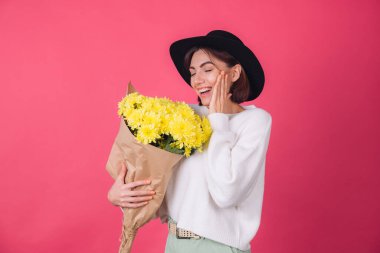 Şapkalı şık bir kadın ve pembe arka planda sıradan beyaz bir süveter. Gülümse. Büyük bir buket sarı çiçek, bahar havası, izole edilmiş bir yer.