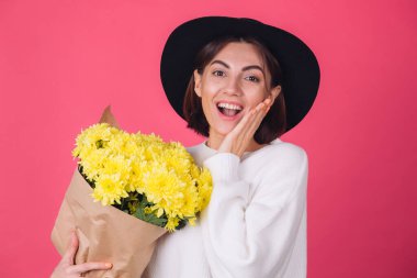 Şapkalı şık bir kadın ve pembe arka planda sıradan beyaz bir süveter. Gülümse. Büyük bir buket sarı çiçek, bahar havası, izole edilmiş bir yer.