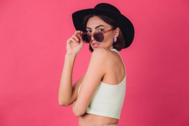 Şapkalı ve güneş gözlüklü güzel bir kadın pembe arka planda poz veriyor.