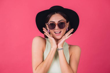 Şapkalı ve güneş gözlüklü güzel bir kadın pembe arka planda poz veriyor. Heyecan verici duygular, izole edilmiş kopya alanı.