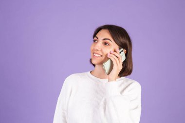 Mor arka planda izole edilmiş beyaz kazaklı genç esmer, mutlu ruh haliyle sohbet ediyor, cep telefonuyla olumlu konuşuyor, gülümsüyor.