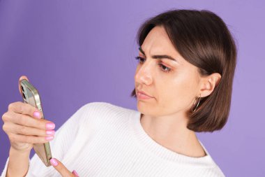 Mor arka planda izole edilmiş beyaz kazaklı genç esmer. Cep telefonlu. Mutsuz suratlı ekrana bak.