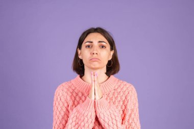 Pembe kazaklı genç esmer, mor arka planda izole edilmiş. Elleriyle birlikte dua ediyor. Yüzünde umut ifadesi var.