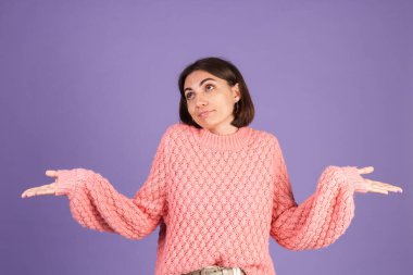 Pembe kazaklı genç esmer, mor arka planda tek başına omuzlarını silkiyor. Hiçbir ipucu yok.