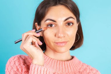 Pembe kazaklı genç esmer, mavi arka planda izole edilmiş. Yüzü makyajlı. Yüzü kızartılmış.