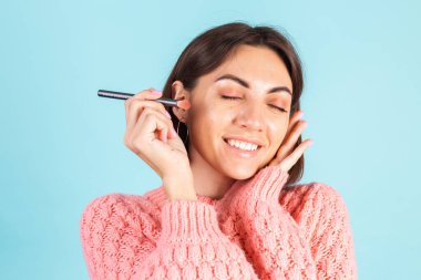 Pembe kazaklı genç esmer, mavi arka planda izole edilmiş. Yüzü makyajlı. Yüzü kızartılmış.