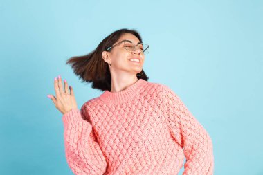 Pembe süveterli genç esmer, mavi arka planda izole edilmiş, gülümseyen gözlükler takıyor.