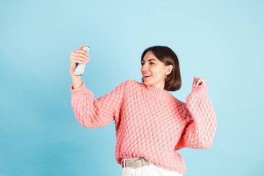 Pembe kazaklı genç esmer, mavi arka planda izole edilmiş gülümsüyor ve selfie çekerken kameraya bakıyor.