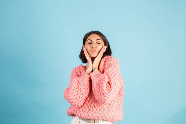 Young brunette in warm pink sweater isolated on blue background happy positive send air kiss to camera