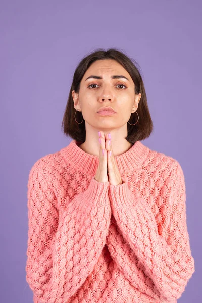 Young brunette in pink sweater isolated on purple background praying with hands together with hope expression on face very emotional and worried