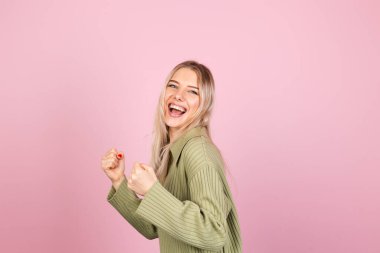 Pembe arka planda sıradan örülü süveter giyen güzel Avrupalı kadın yumruğunu sıkarak kutlama yapıyor.