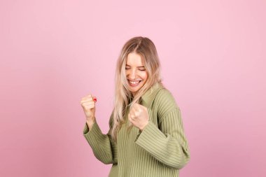 Pembe arka planda sıradan örülü süveter giyen güzel Avrupalı kadın yumruğunu sıkarak kutlama yapıyor.