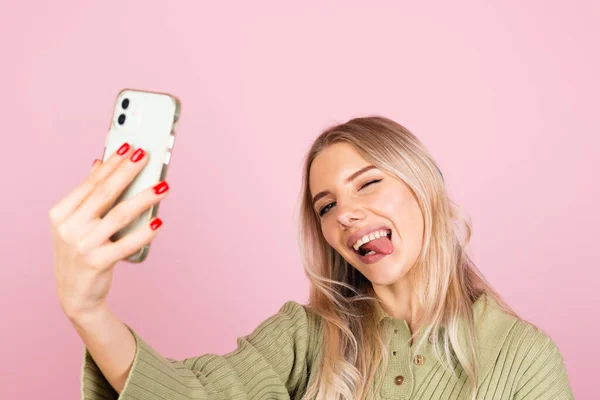 Pretty european woman in casual knitted sweater on pink background taking selfie photo on phone does funny grimaces shows tongue positive
