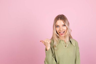 Pembe arka planda gündelik süveter giyen güzel Avrupalı bir kadın baş parmağıyla mutlu bir gülümseme noktası bırakıyor.