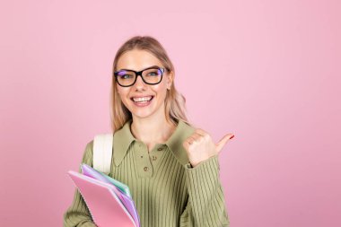 Pembe arka planda sıradan süveter giyen güzel Avrupalı bir kadın. Gülümseyen defterler, mutlu parmak gösterme, sağ eğitim kavramı.