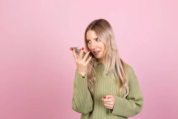 ピンクの背景にカジュアルなセーターのかなりヨーロッパの女性は 携帯電話の録音音声メッセージ幸せな笑顔を保持し 会話を持ち 陽気に興奮 — ストック写真