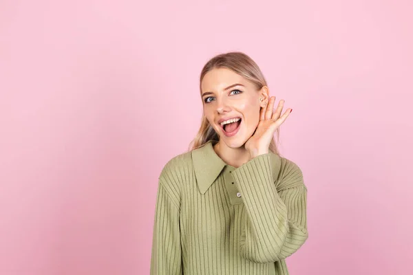 かなりヨーロッパの女性でカジュアルなセーターでピンクの背景好奇心は あなたが耳のコピースペースで手で何を言って聞くしようとする — ストック写真