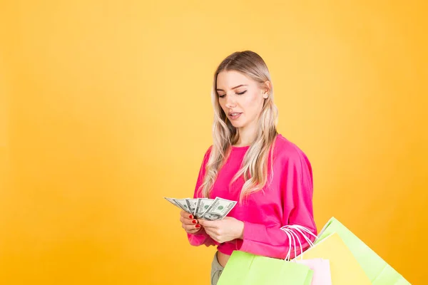 Pretty european woman in pink  blouse on yellow background smile excited hold fan of 100 dollars money and shopping bags count money
