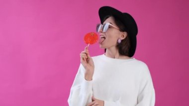 Pembe arka planda beyaz süveterli ve şapkalı bir kadın izole edilmiş büyük turuncu bir lolipop yalıyor.  
