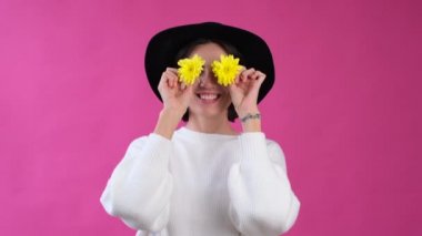 Pembe arka planda beyaz süveterli ve şapkalı bir kadın izole edilmiş mutlu, gülümseyen, iki sarı çiçekli gözler.