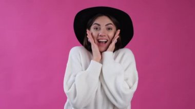 Pembe arka planda rahat beyaz kazaklı ve şapkalı bir kadın kameraya bakıyor ve şaşkın şaşkın bir yüz olduğunu söylüyor. 