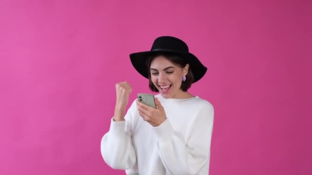 女性でカジュアルホワイトセーターと帽子オンピンクの背景絶縁幸せとともに携帯電話行いますWinジェスチャークリーニング拳 — ストック動画