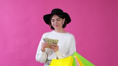 Sıradan beyaz kazaklı ve pembe şapkalı bir kadın alışveriş torbaları ve 100 dolarlık para sayma hayranıyla izole edilmiş.  