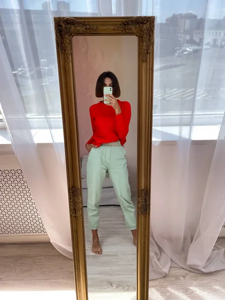 在家里穿着鲜艳的红色毛衣和薄荷绿色裤子的时尚女性在手机上拍照 这是在手机上的社交媒体上拍摄的 — 图库照片