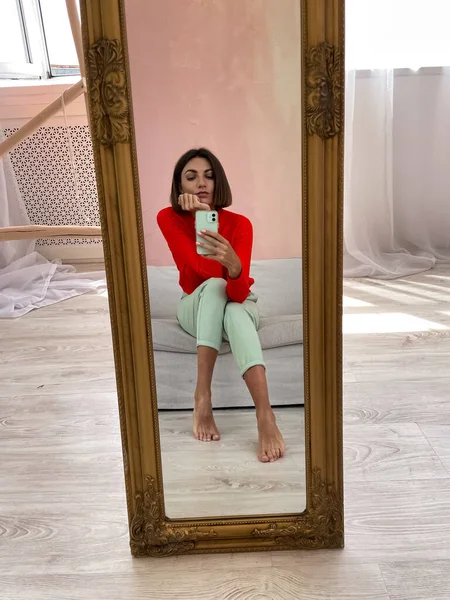 明るい赤いセーターとミントグリーンのパンツで自宅でスタイリッシュな女性は物語のための電話で鏡の中で写真を撮るソーシャルメディア — ストック写真