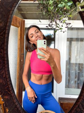 Evdeki güzel kadın cep telefonundaki aynada fotoğraf çekiniyor sosyal medyadaki haber ve paylaşımlar için, dikey çerçeve, spor kıyafetleri, forma ve ince, karın kasları motivasyonu