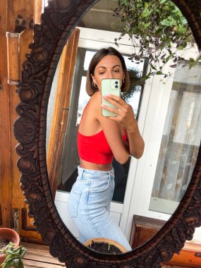 Evdeki güzel kadın cep telefonundaki fotoğrafların aynada fotoğrafını çekiyor sosyal medya, dikey çerçeve, kot pantolon ve kırmızı bluz giyiyor, ince, güzel görünümlü.